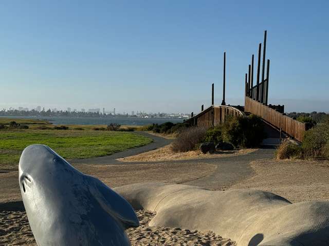 Sculpture of a whale on front left w/ a path wooden observation platform to the right going a little further back. Water and tall buildings are in the background.